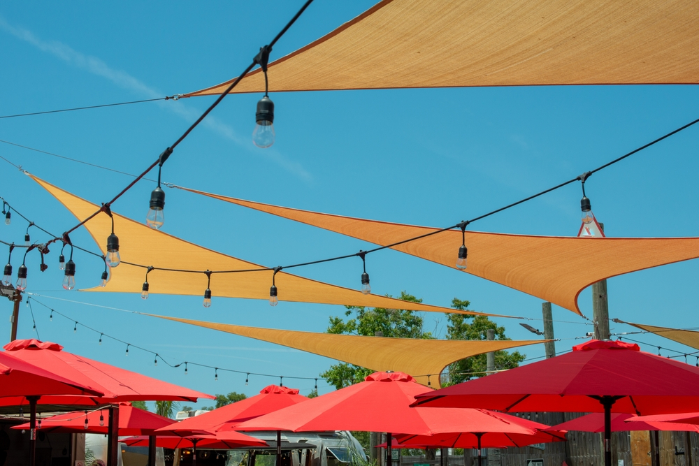 Multiple triangle shaped yellow nylon sunshades and awnings hanging over a patio deck. There are red colored canvas umbrellas hung with strings of clear patio light against a bright blue sunny sky.